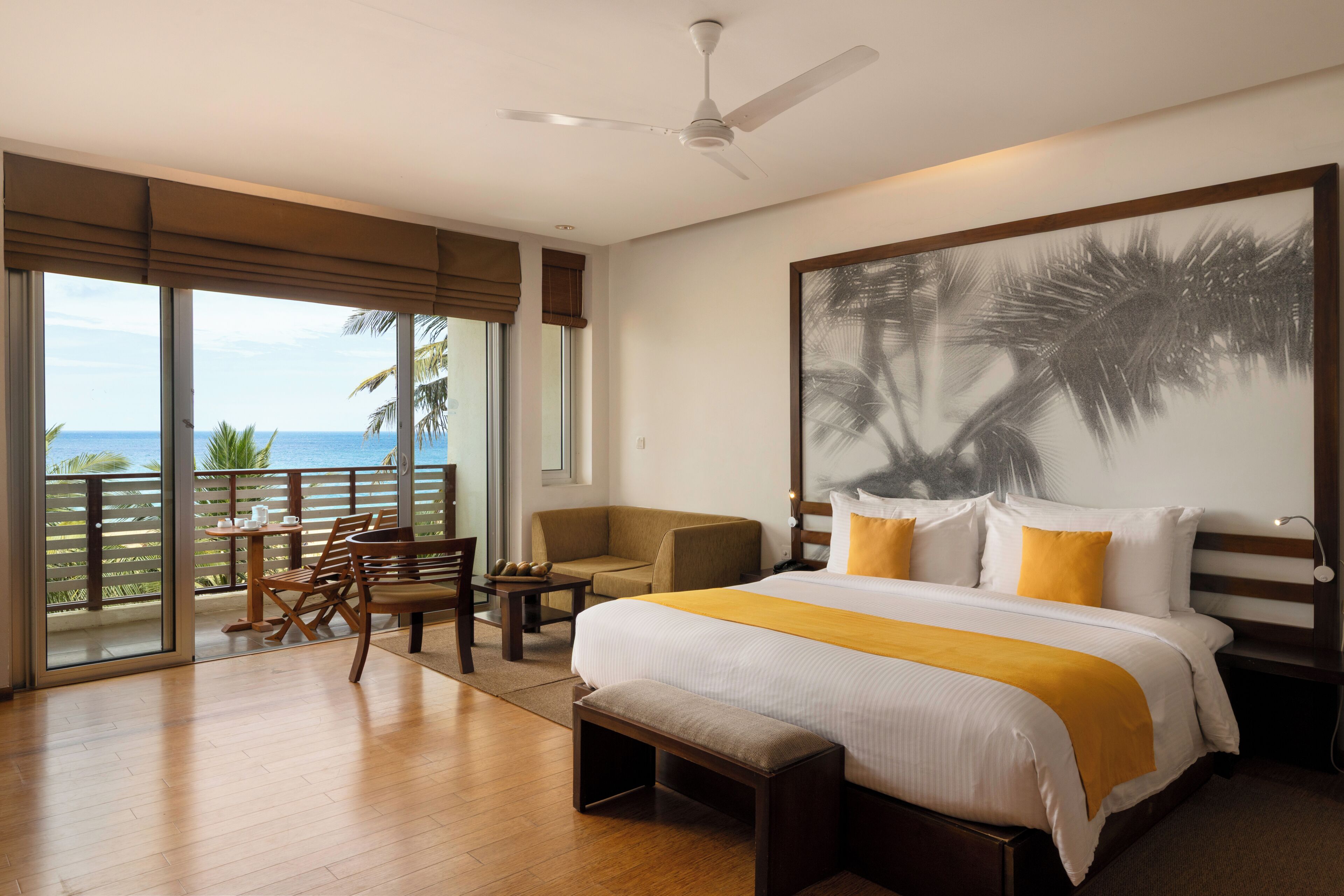 A hotel room with a large bed, a ceiling fan, a balcony with a view of the ocean, and a painting of palm trees.
