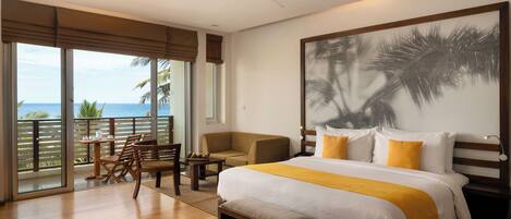 Habitación de hotel con una cama grande, ventilador de techo, balcón con vista al océano y un cuadro de palmeras.