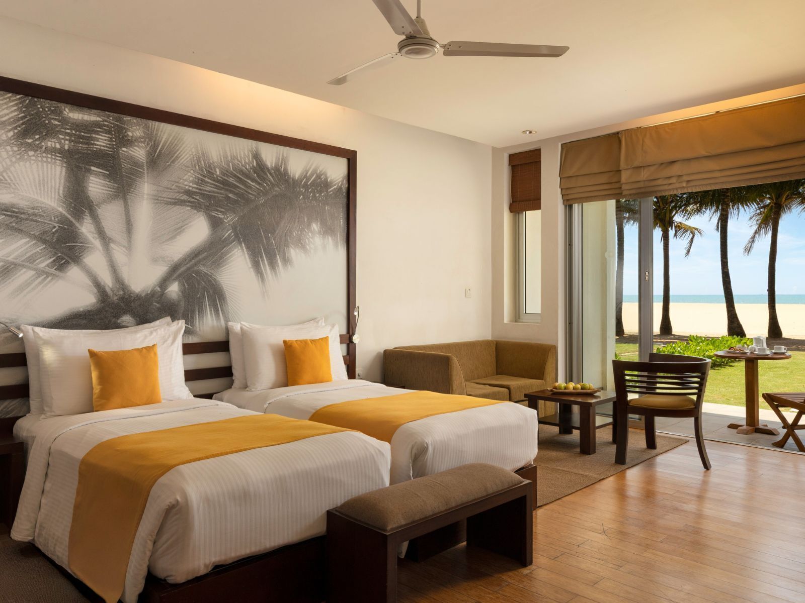 Habitación de hotel con dos camas, un banco, una silla, una mesa con frutas y vista a la playa.