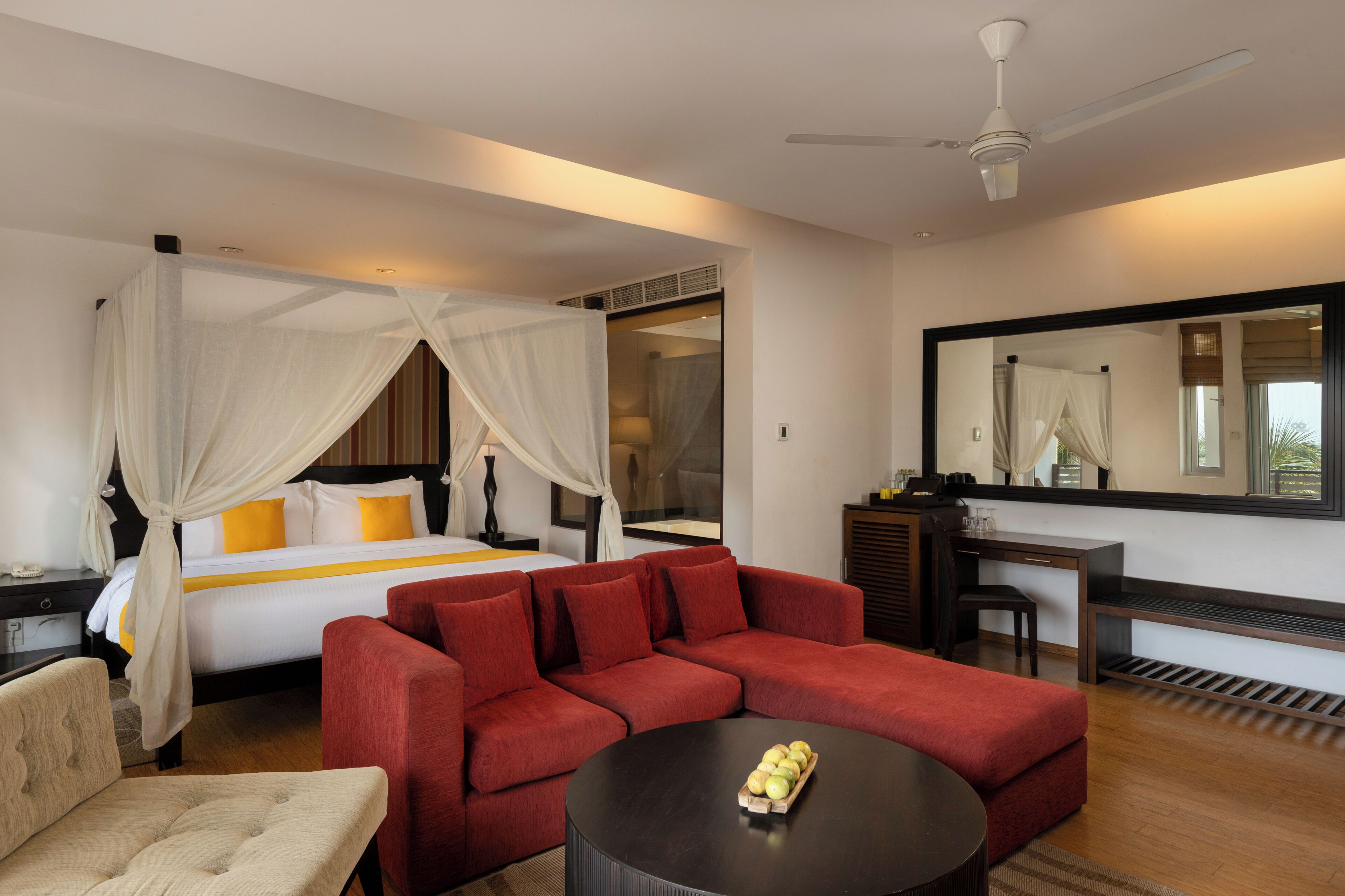 A hotel room with a bed, a red sofa, a round coffee table, a desk, and a large mirror.