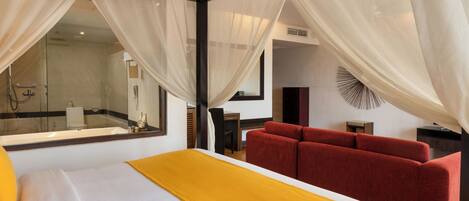 Habitación de hotel con cama, sofá rojo, espejo grande y vista al baño.