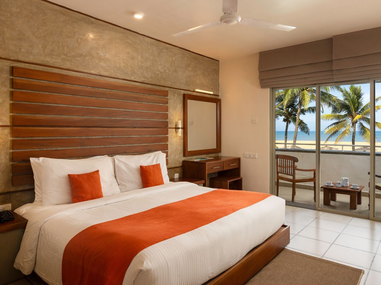 Een hotelkamer met een groot bed, een bureau, een stoel en een balkon met uitzicht op het strand en palmbomen.
