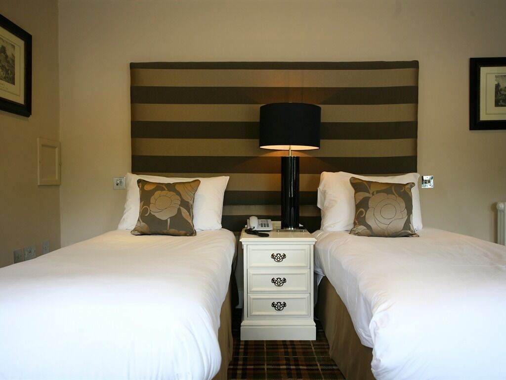 Quarto de hotel com duas camas, cabeceira com listras horizontais, luminária de cabeceira e uma pequena mesa de cabeceira branca com gavetas.
