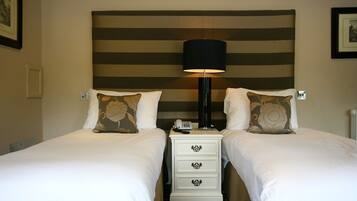 Quarto de hotel com duas camas, cabeceira com listras horizontais, luminária de cabeceira e uma pequena mesa de cabeceira branca com gavetas.
