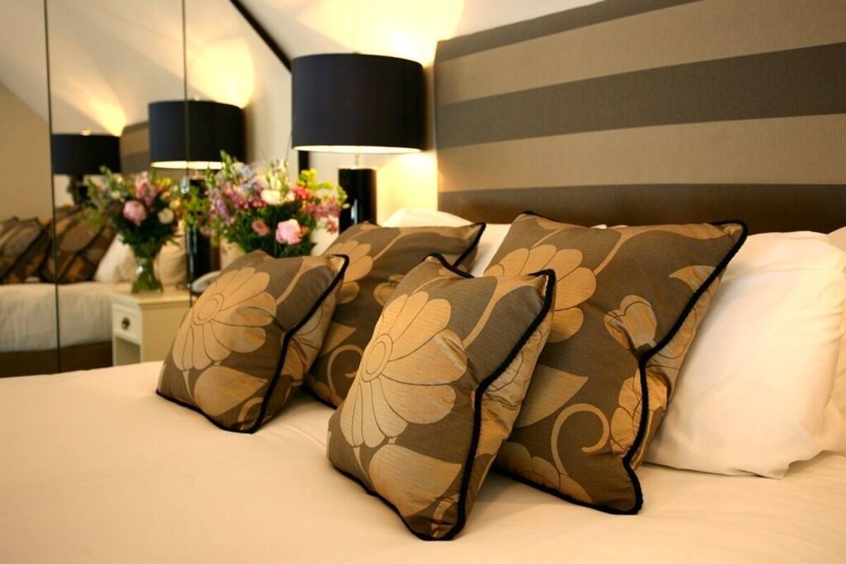 Une chambre d’hôtel avec un lit, des oreillers à motifs floraux, une table de chevet avec une lampe et un miroir qui reflète la pièce.
