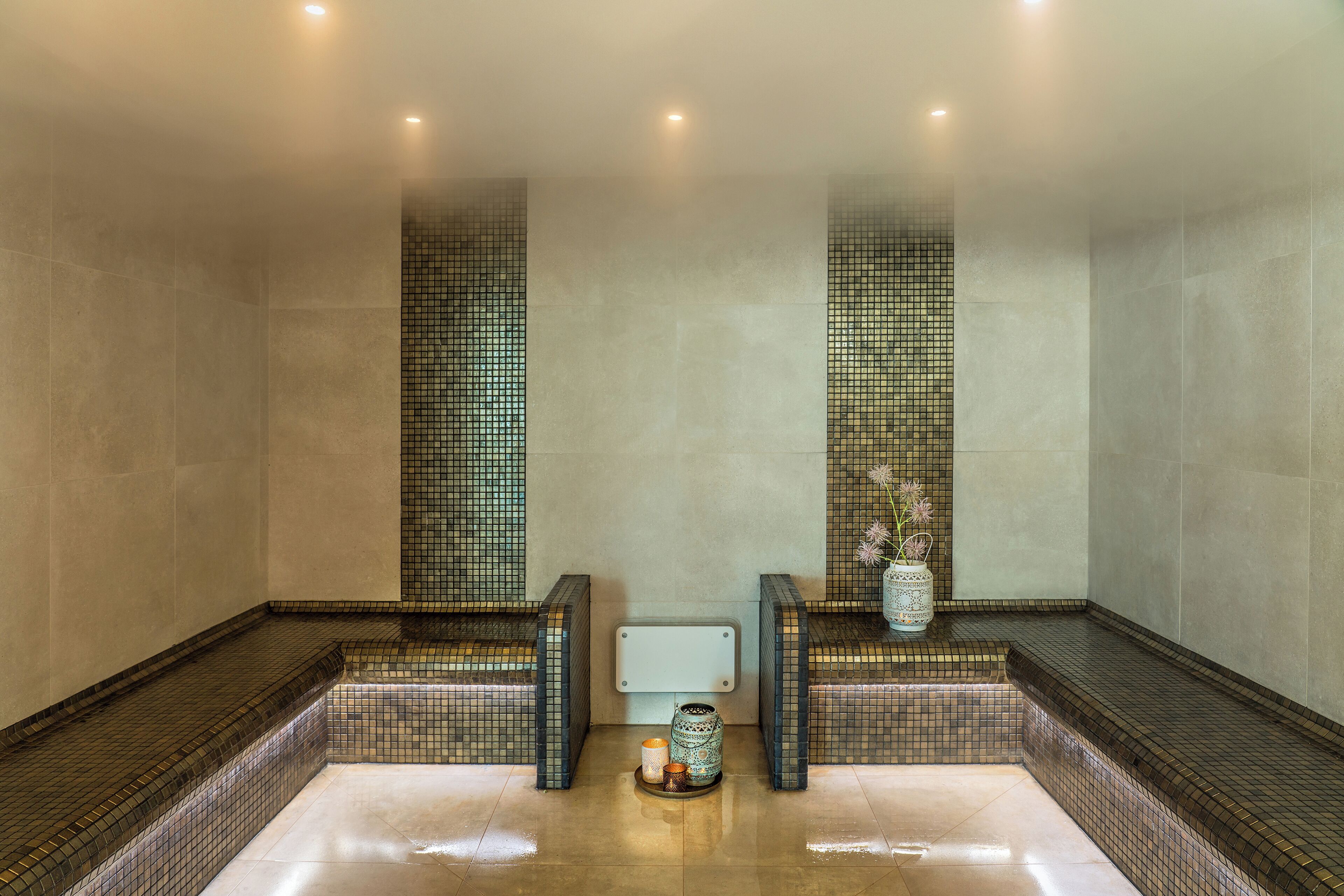 A spa area with a tiled floor, two benches, a small table with a candle and a vase, and a wall-mounted light fixture.