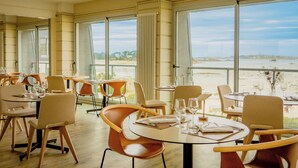 Un comedor con mesas y sillas, ventanas amplias con vista a la playa y un mar tranquilo.
