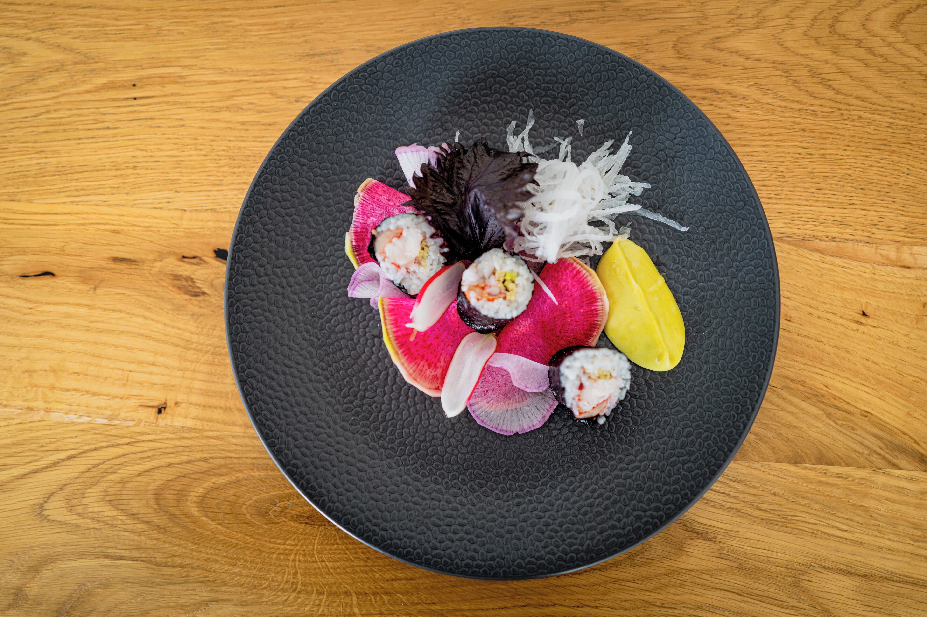 Une assiette noire ronde, sur laquelle sont disposés différents morceaux de sushi, est posée sur une table en bois.