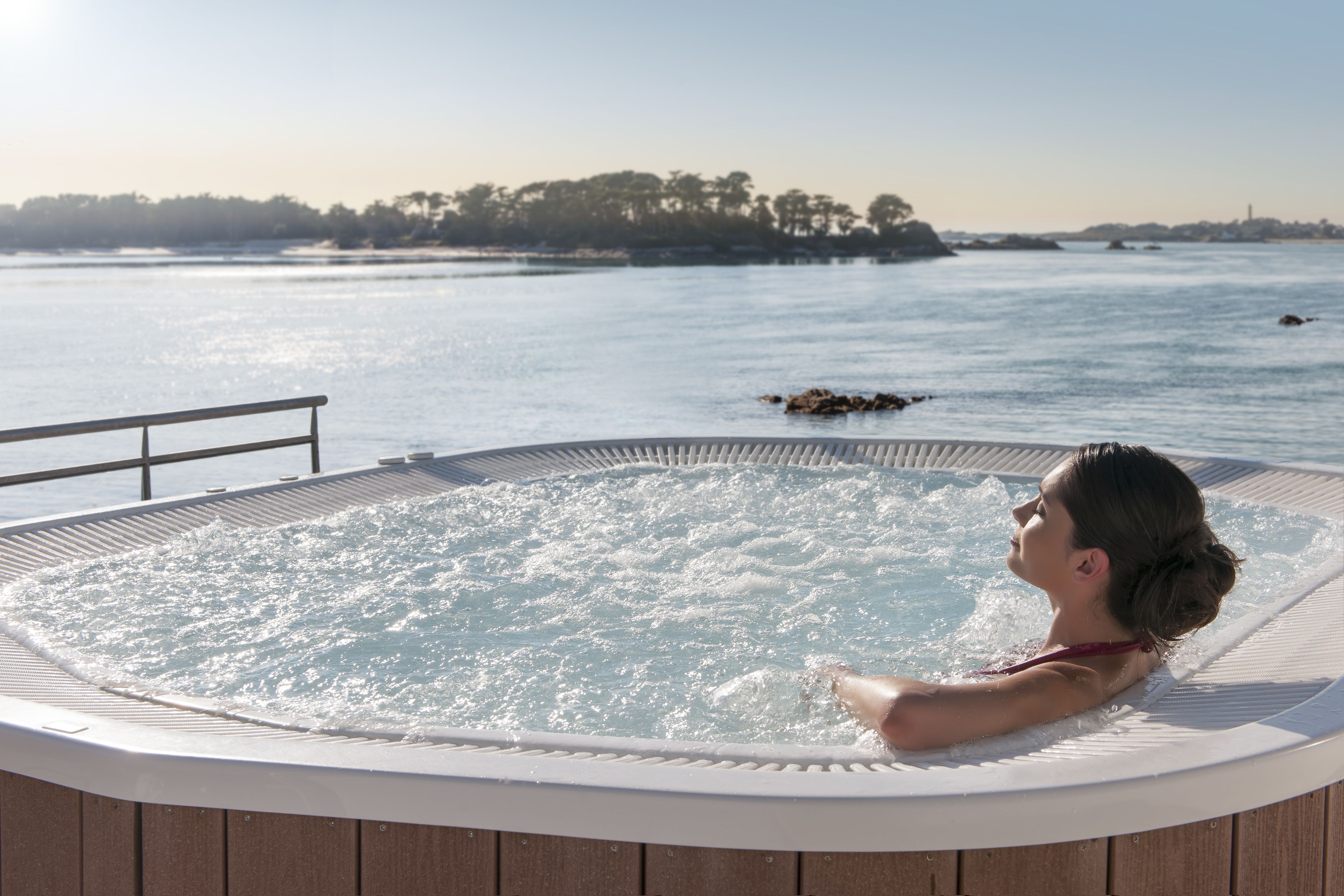 A hot tub with a wooden deck, overlooking a body of water with a distant shoreline.