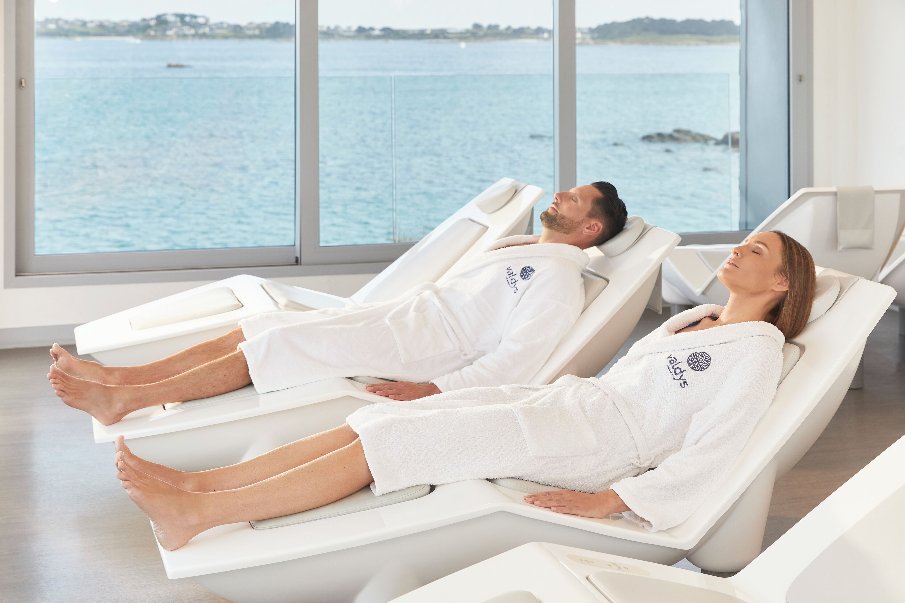 Two individuals lying on separate reclining chairs in a room with large windows overlooking a body of water.