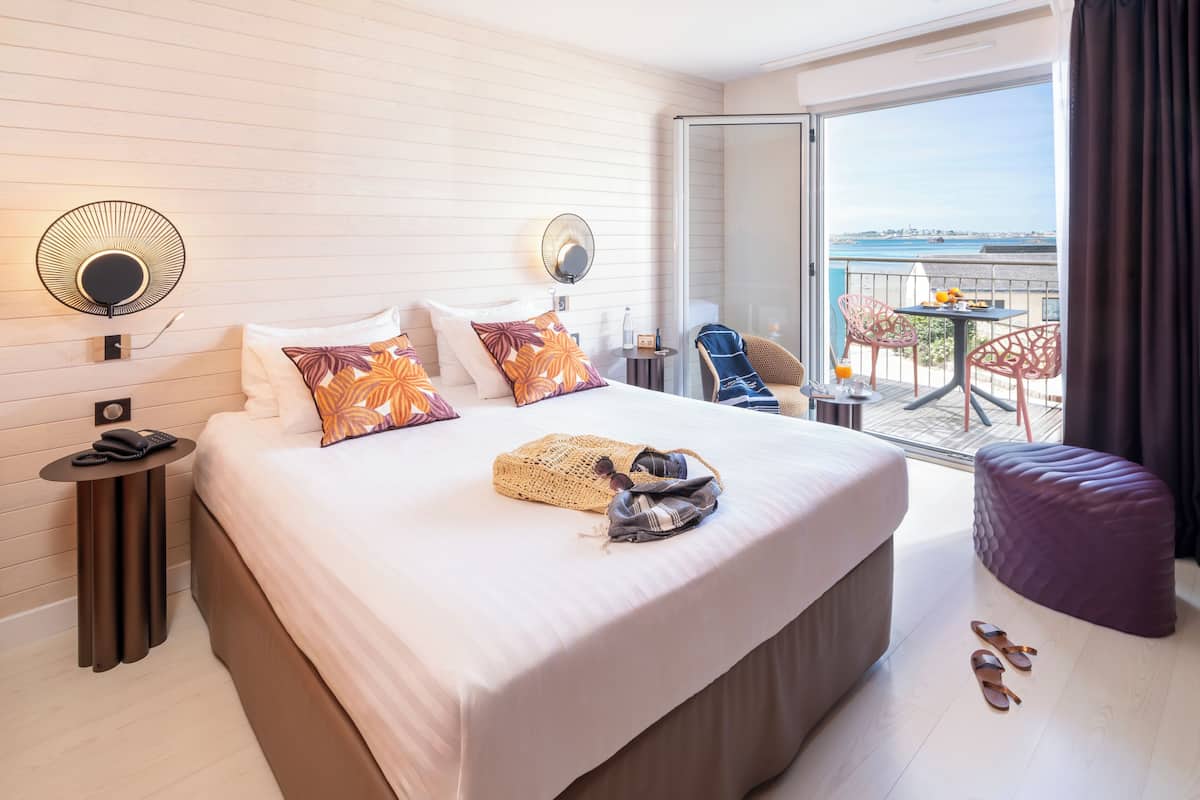 Een hotelkamer met een groot bed, een klein tafeltje met een stoel, een balkon met een tafel en stoelen, en uitzicht op zee.