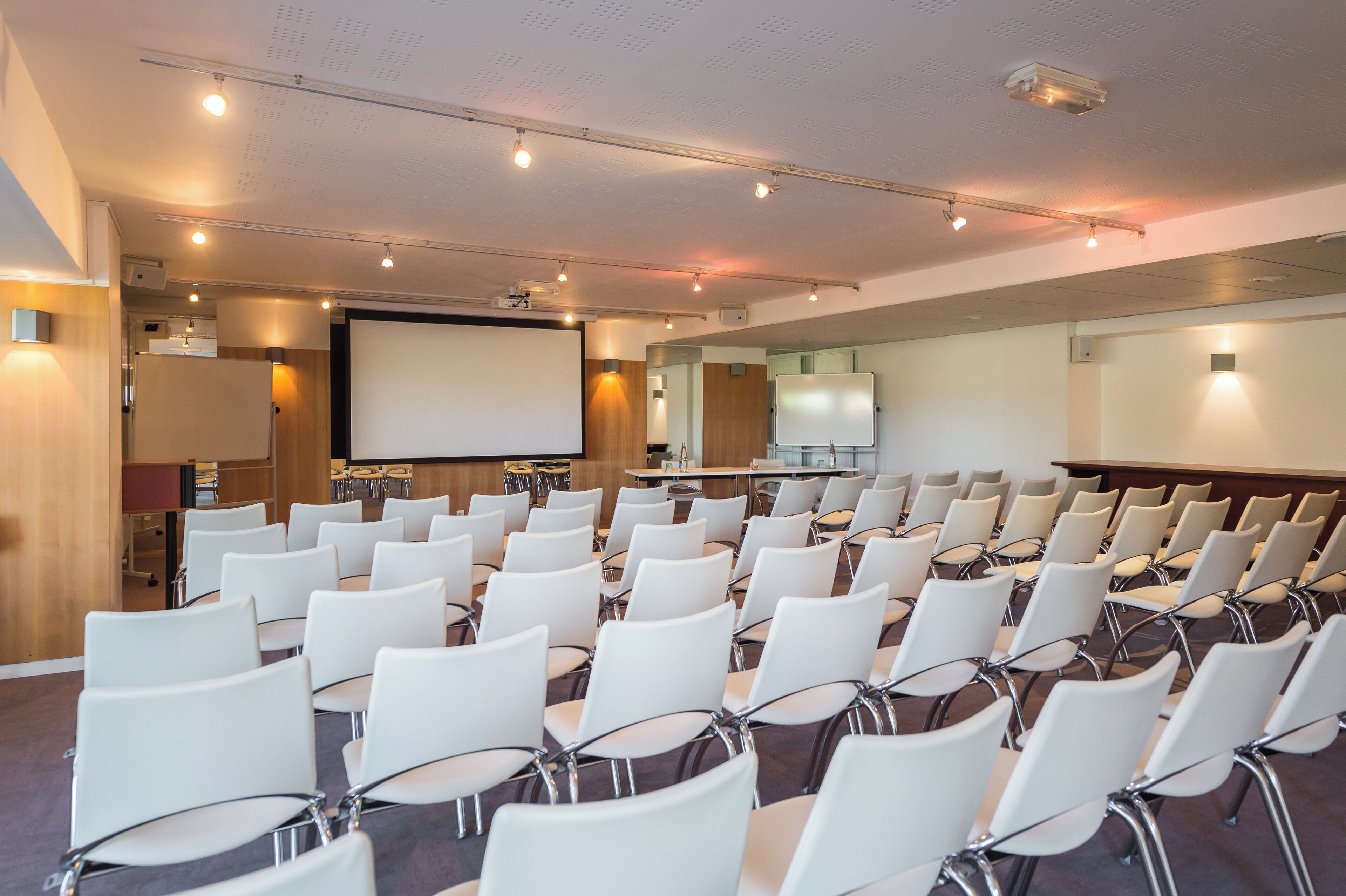 Een vergaderruimte met rijen witte stoelen, een projectiescherm en een whiteboard.