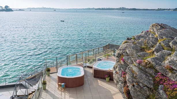 Una terraza con dos jacuzzis que dan a una masa de agua, una escalera y terreno rocoso con vegetación.