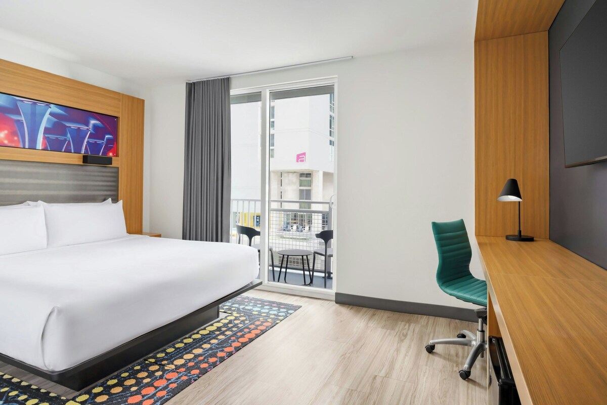 A hotel room with a bed, a desk, a chair, a TV, and a view of the outside.
