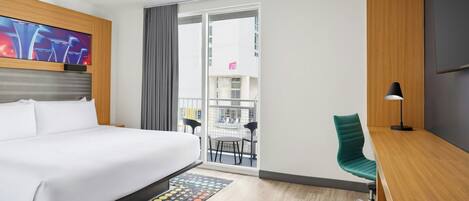A hotel room with a bed, a desk, a chair, a TV, and a view of the outside.
