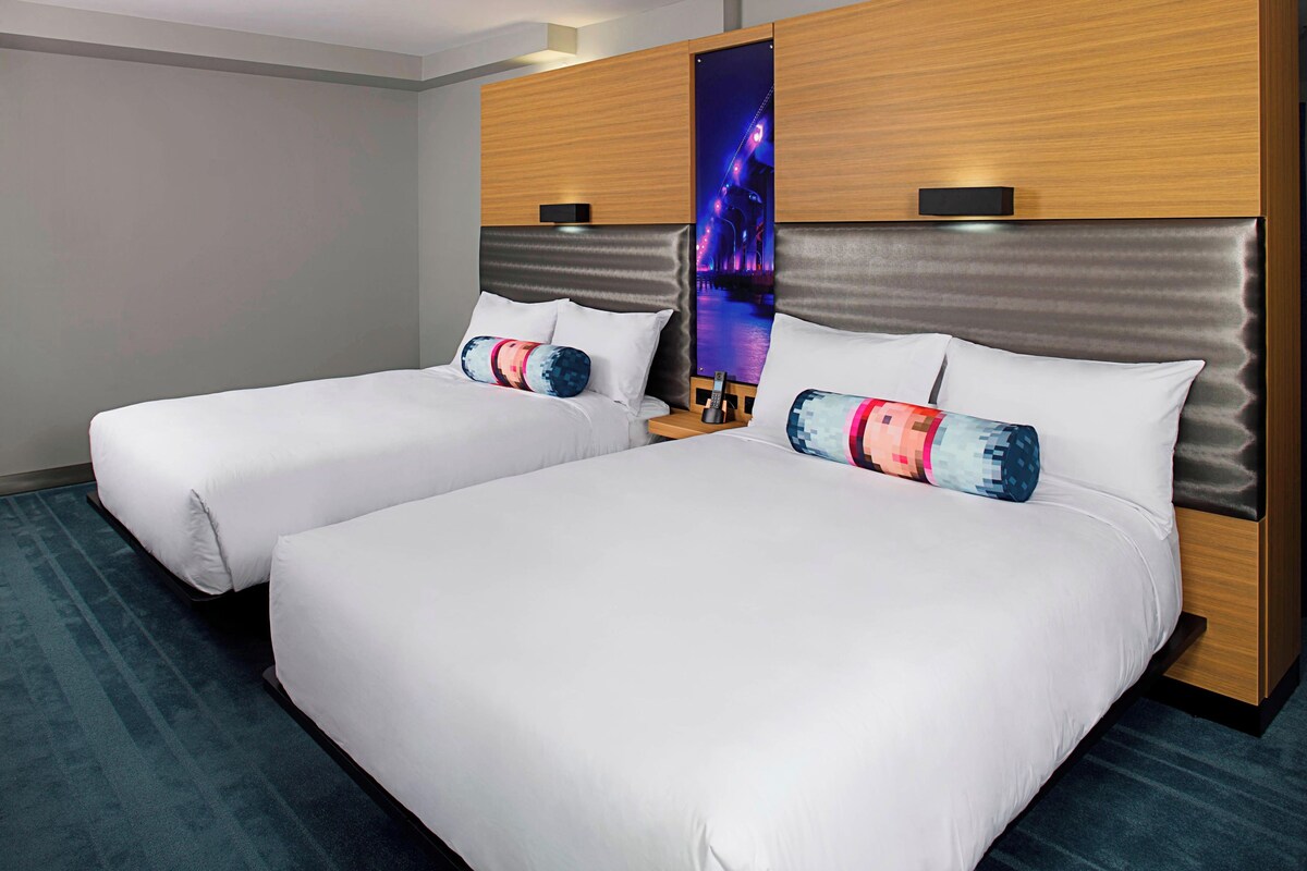 Habitación de hotel con dos camas, una mesita de noche, televisor montado en la pared y vista panorámica de la ciudad.