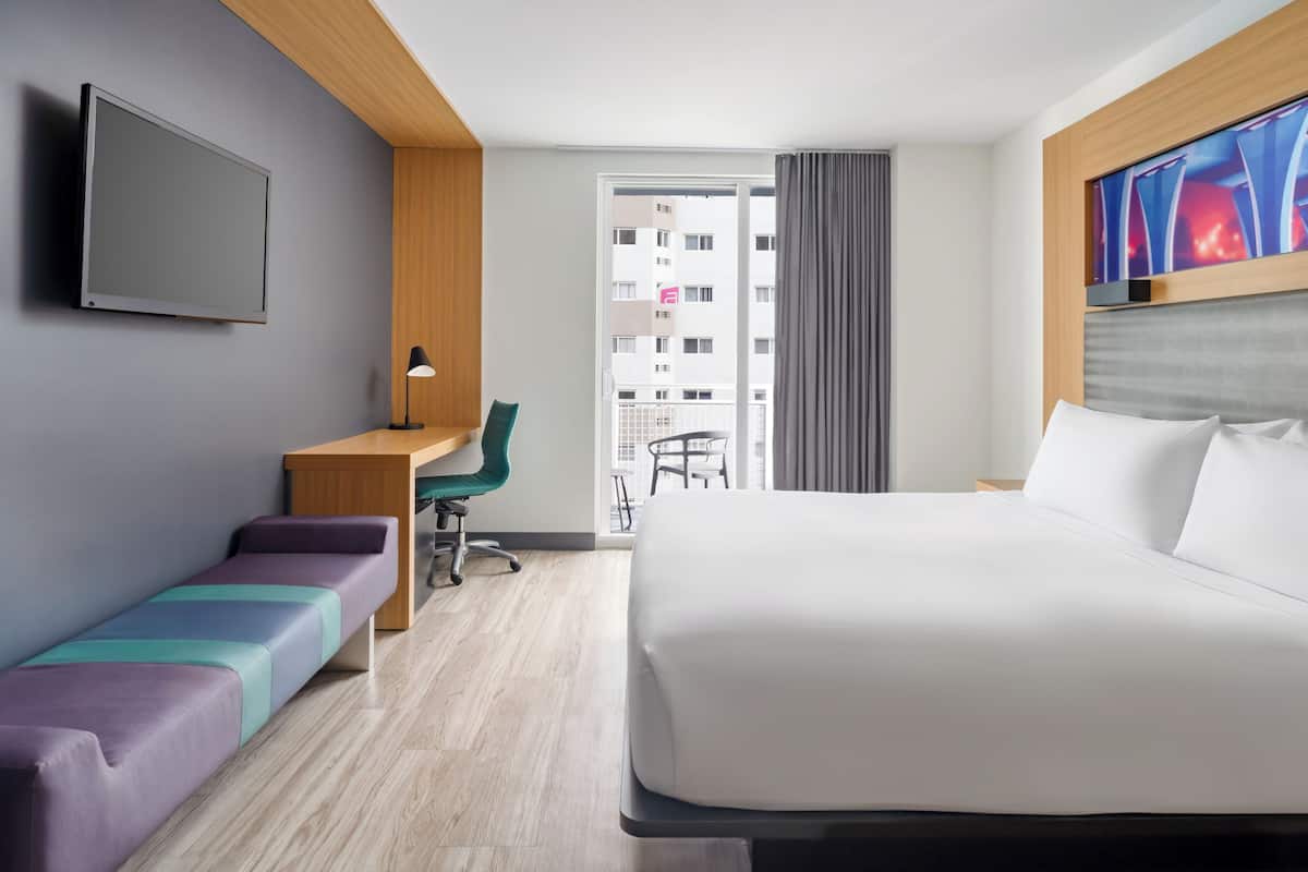A hotel room with a bed, a desk, a chair, a bench, a TV, and a view of the outside.