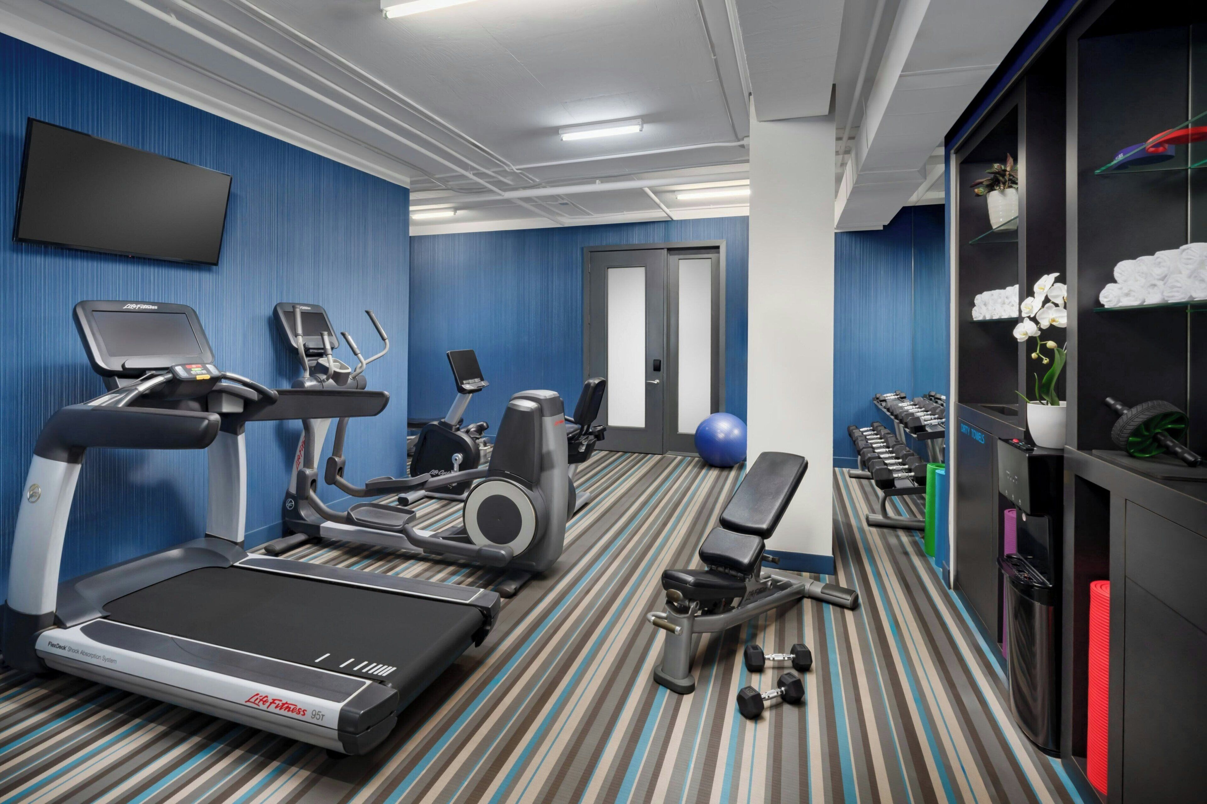 A hotel fitness center with cardio machines, dumbbells, and a yoga mat.