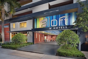 Una entrada de hotel moderna con un letrero colorido, un corredor cubierto y jardines bien cuidados.