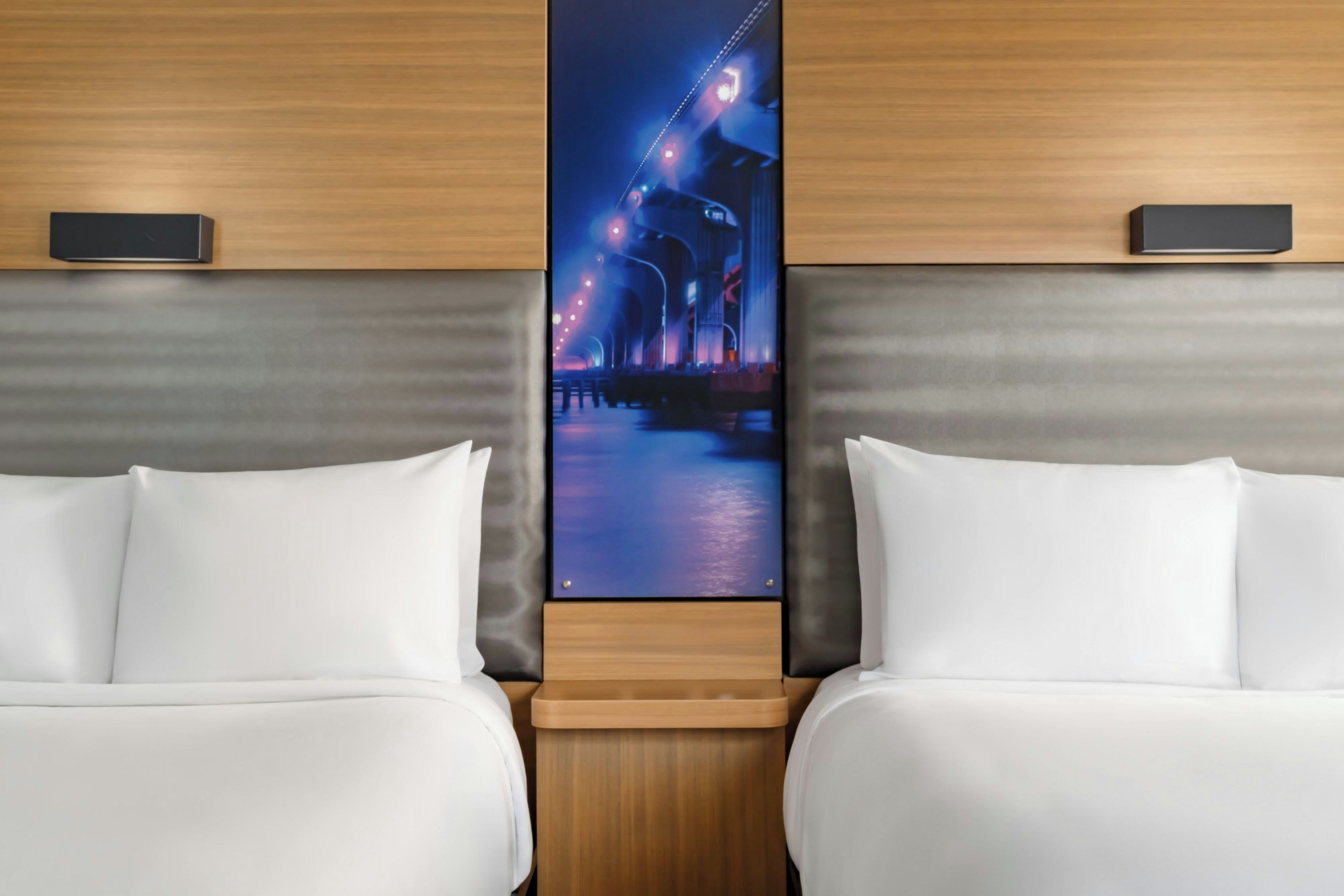 A hotel room with two beds, a large wall-mounted TV displaying a cityscape, and a wooden headboard.