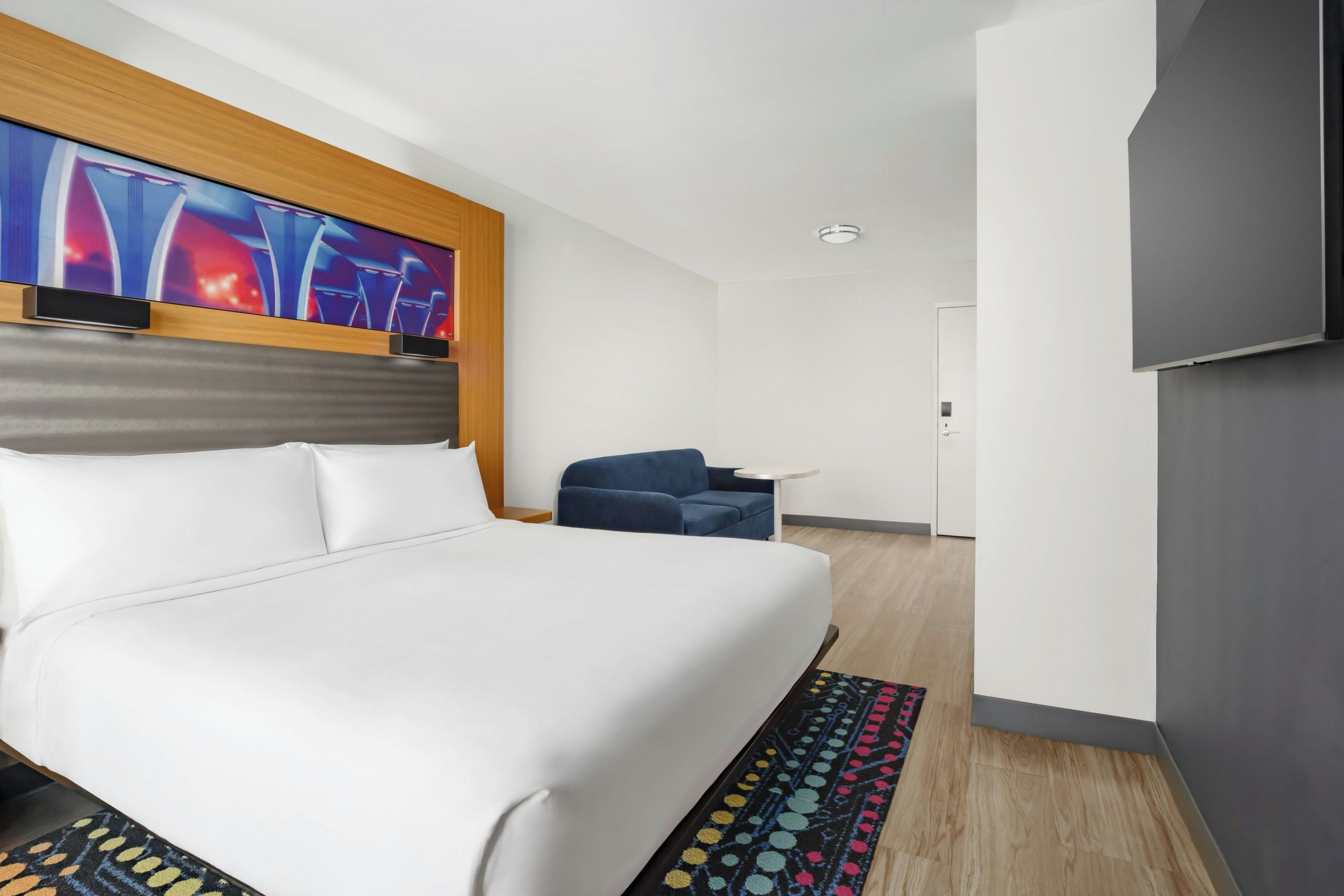 Habitació d'hotel amb un llit gran, televisió penjada a la paret, taula petita i una porta.