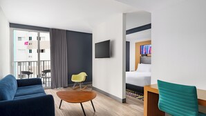 Una habitación de hotel moderna con sofá, una mesita redonda pequeña, un escritorio de madera, una silla color turquesa, televisor montado en la pared y una cama con cabecera.