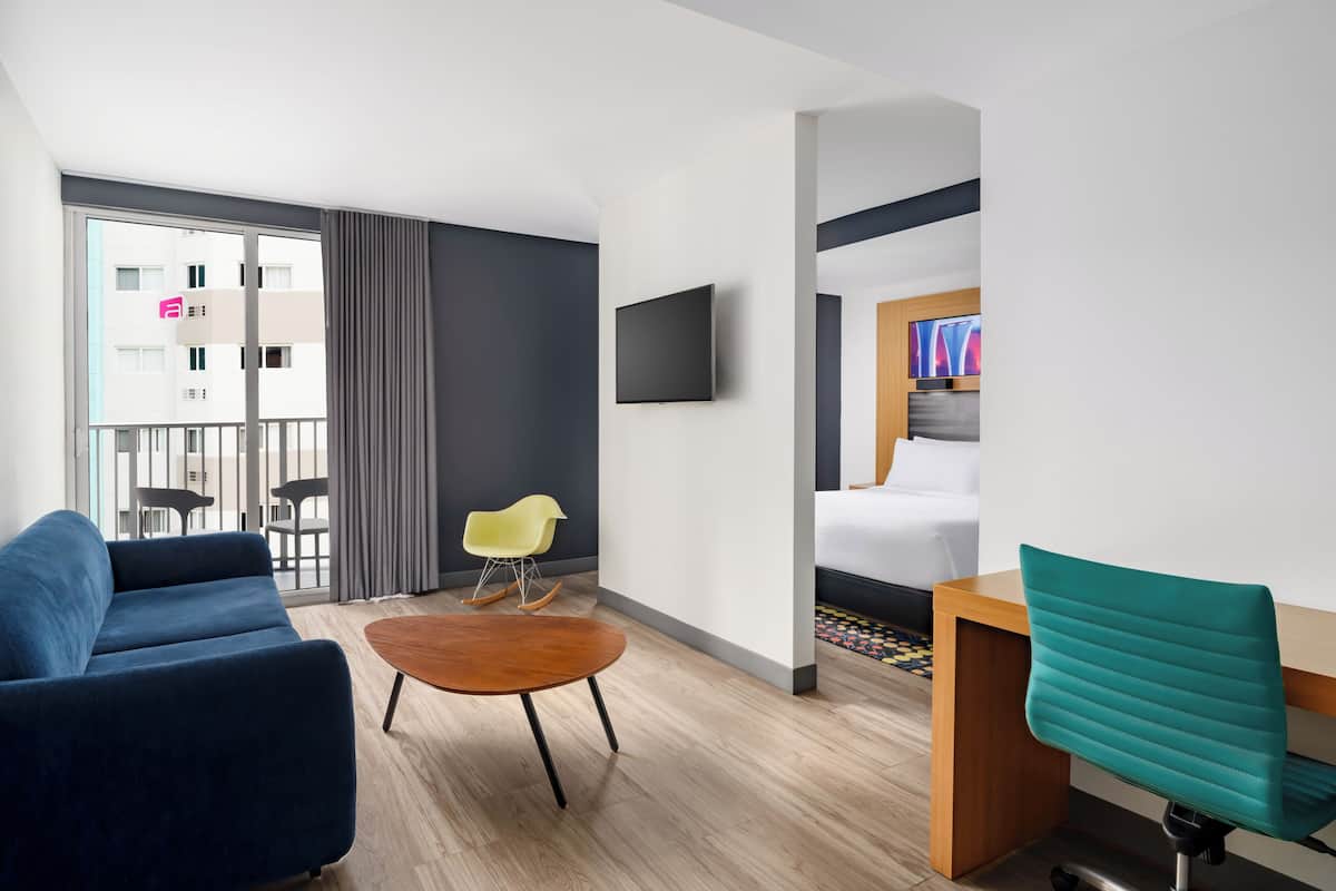 A modern hotel room with a sofa, a small round table, a wooden desk, a teal chair, a TV mounted on the wall, and a bed with a headboard.