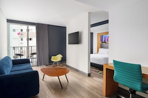 A modern hotel room with a sofa, a small round table, a wooden desk, a teal chair, a TV mounted on the wall, and a bed with a headboard. - Aloft by Marriott Miami - Brickell (Miami)