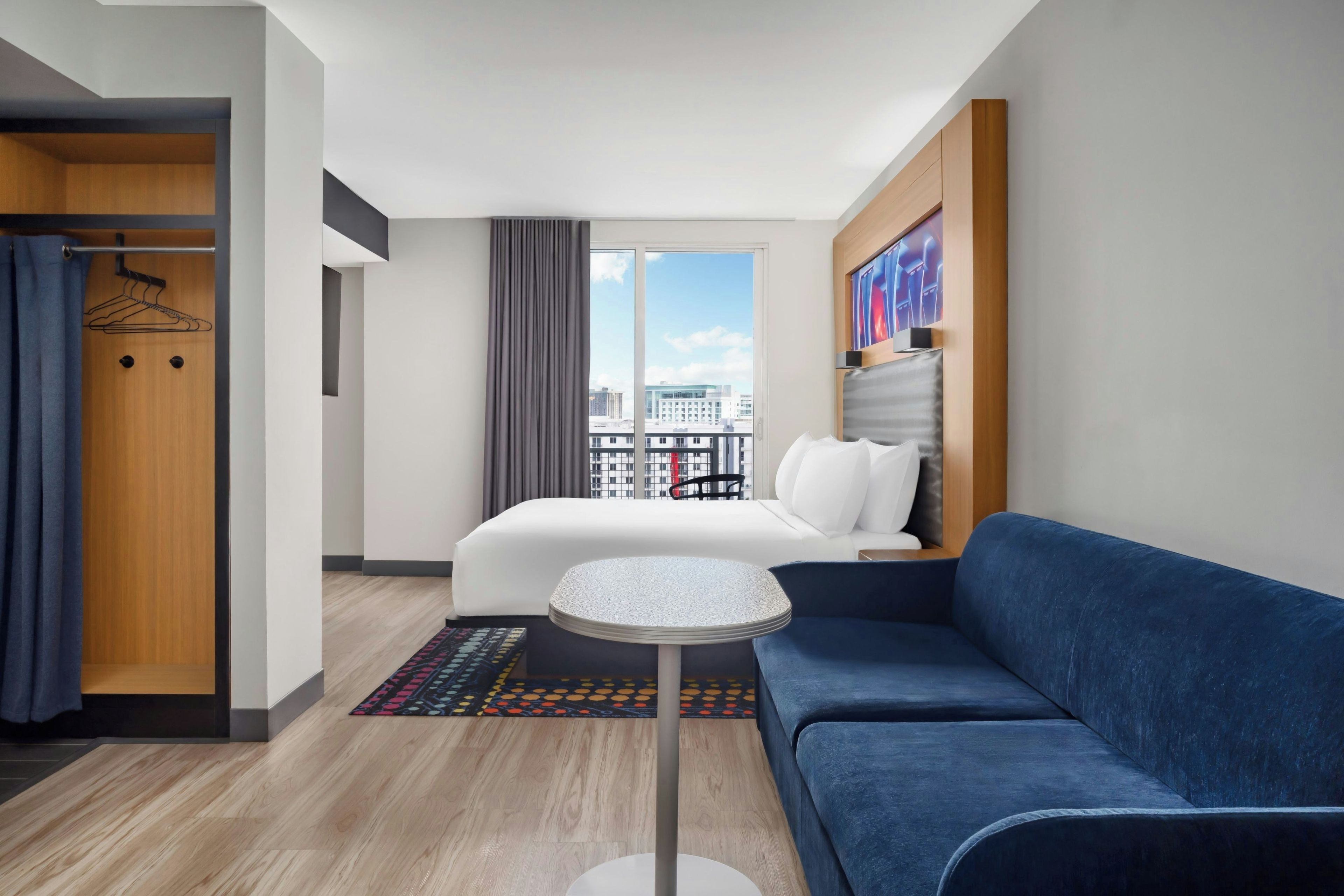 A hotel room with a bed, a blue sofa, a small round table, a wardrobe, and a view of the city.