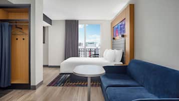A hotel room with a bed, a blue sofa, a small round table, a wardrobe, and a view of the city.