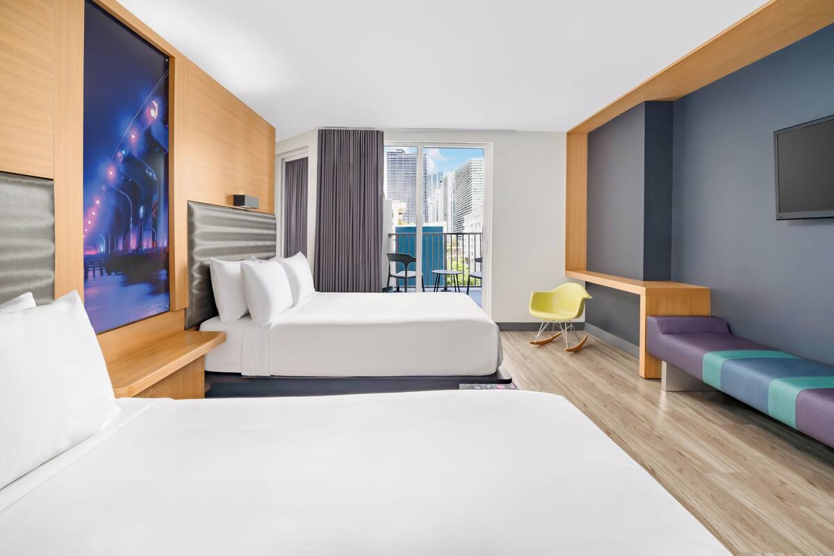 A hotel room with a large bed, a sofa, a TV, a balcony with a view of buildings, and a chair.