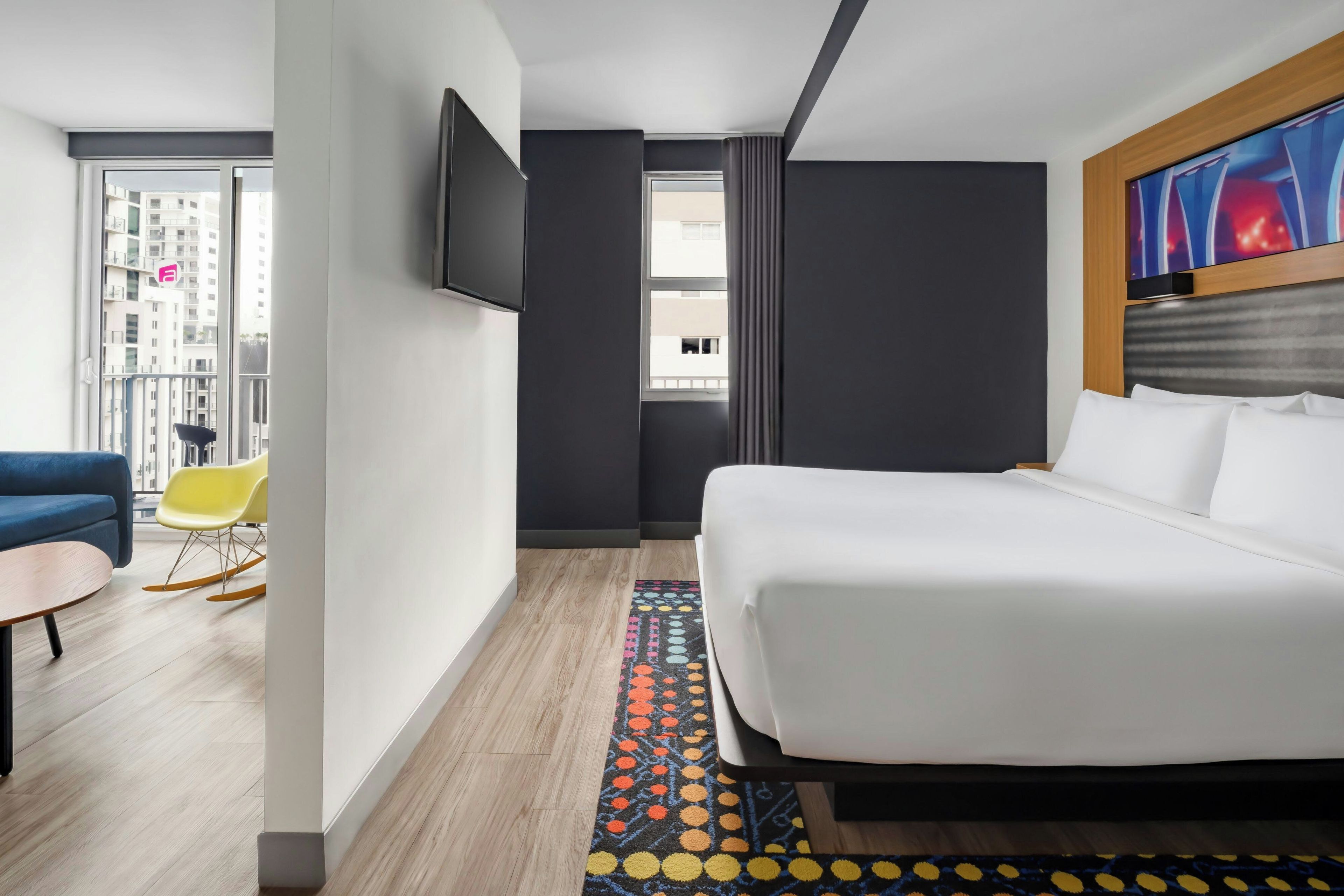 Ein Hotelzimmer mit einem Bett, einem Fernseher, einem Fenster, einer Couch, einem Sessel und einem gemusterten Teppich.