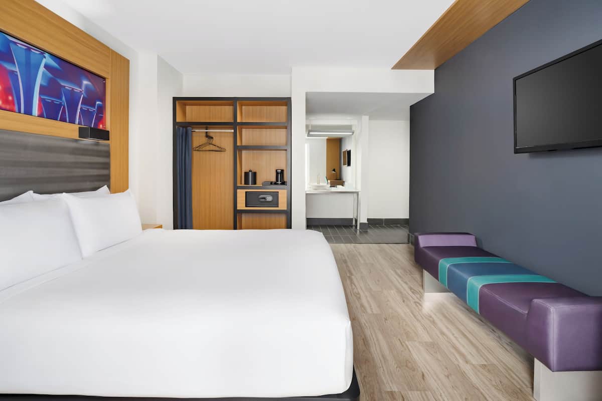 A modern hotel room with a large bed, a walk-in closet, a kitchenette, a seating area with colorful cushions, and a TV mounted on the wall.