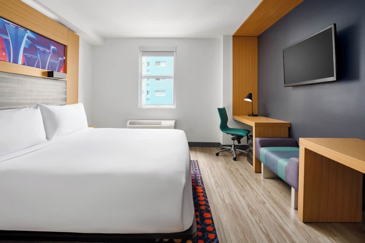 A modern hotel room with a large bed, a desk, a chair, a TV, and a window with a view of buildings.