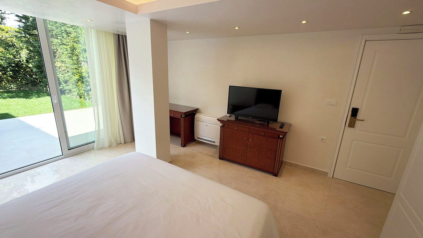 Un dormitorio con una cama grande, un televisor sobre un mueble de madera, un escritorio y un ventanal corredera que da a un balcón.