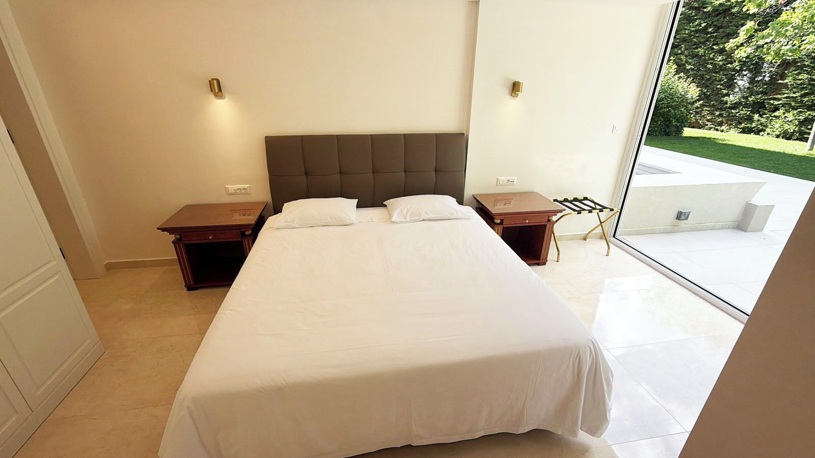 A bedroom with a large bed, two bedside tables, a nightstand, and a view of a garden through a glass door.
