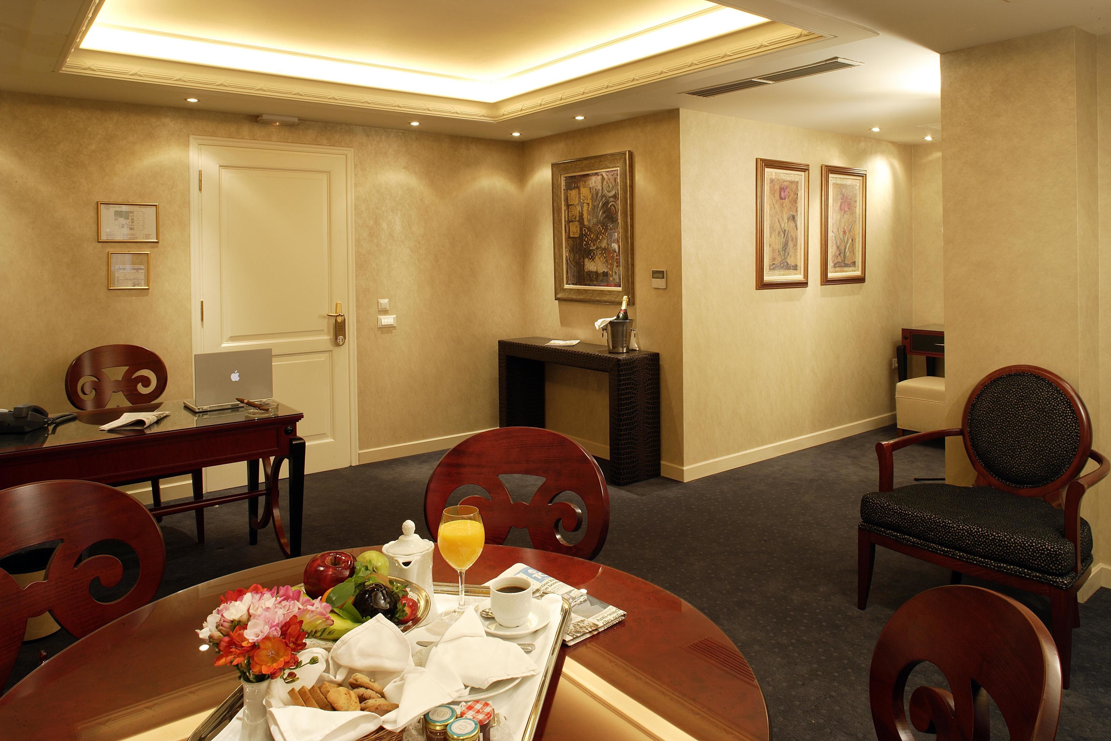 A hotel room with a dining area, a desk with a laptop, a minibar, and a painting on the wall.