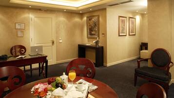 A hotel room with a dining area, a desk with a laptop, a minibar, and a painting on the wall.