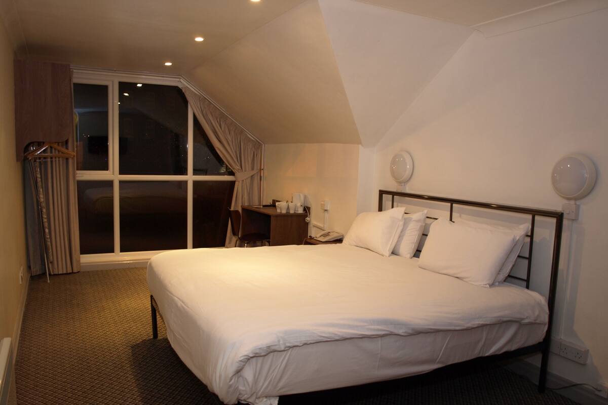 A bedroom with a large bed, a desk, a chair, a window with curtains, and a nightstand with a phone and a lamp.