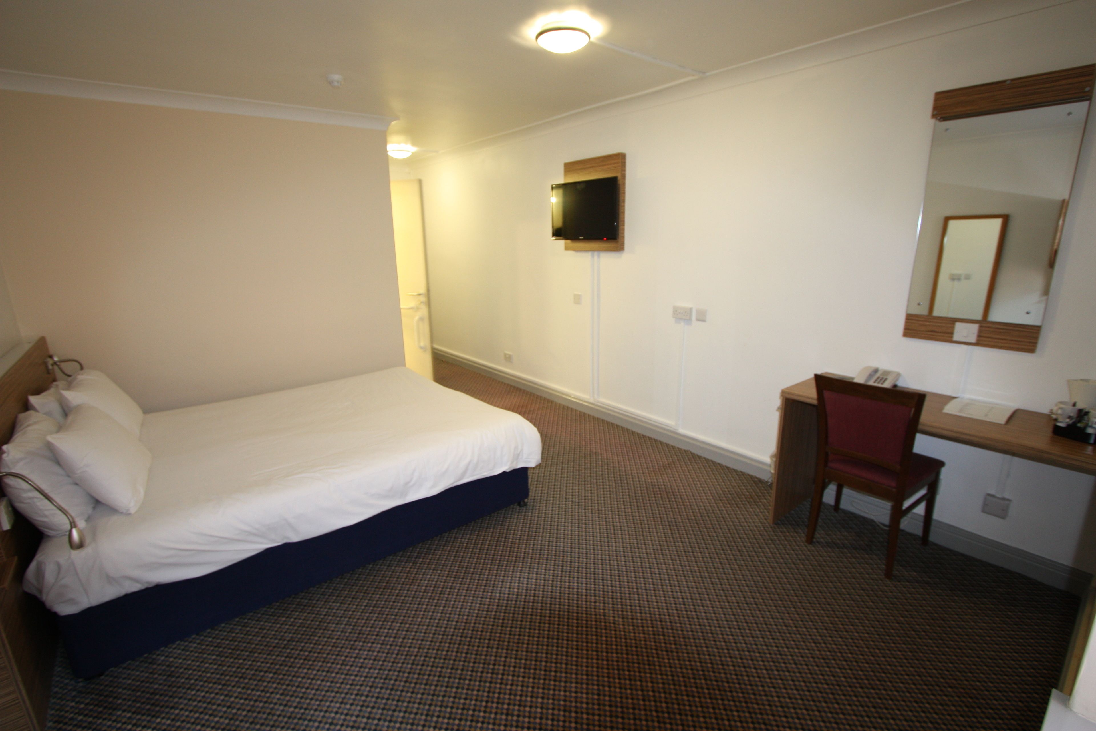 A hotel room with a bed, a desk, a chair, a TV mounted on the wall, and a mirror.