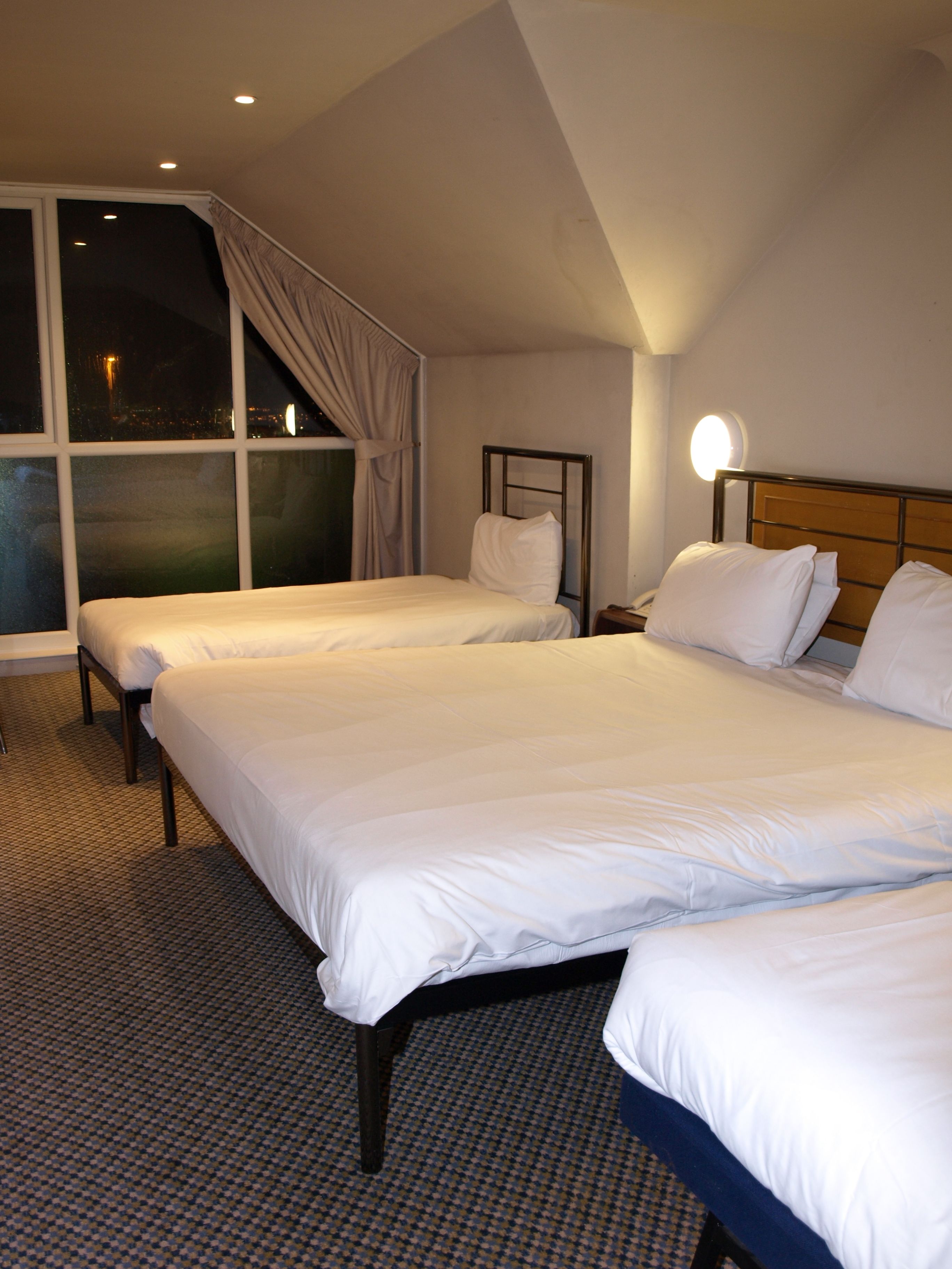 A hotel room with two single beds, a large window with curtains, a bedside table with a lamp, and a checkered floor.