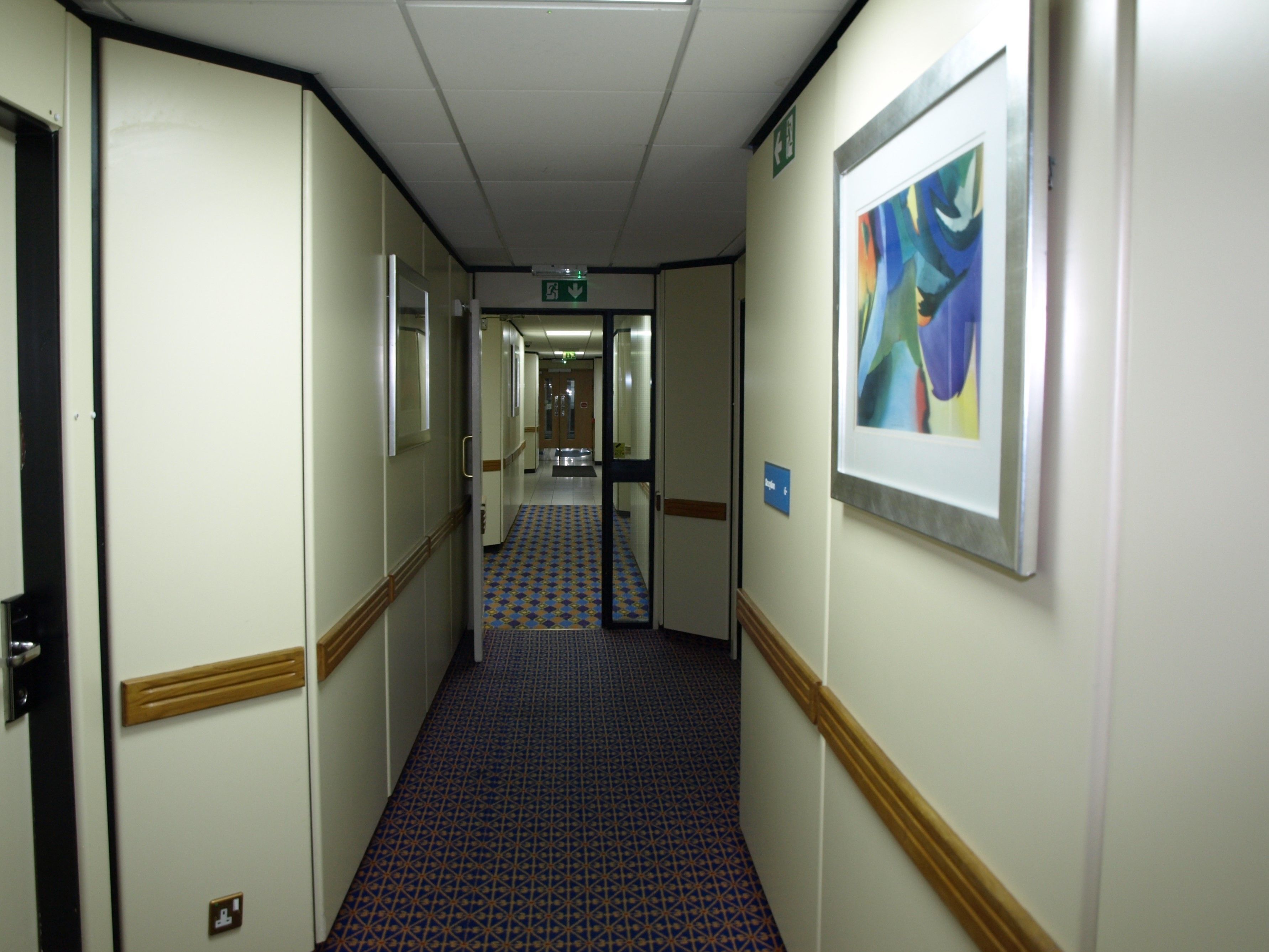 A hotel hallway with patterned carpet, framed artwork on the walls, and doors leading to rooms.