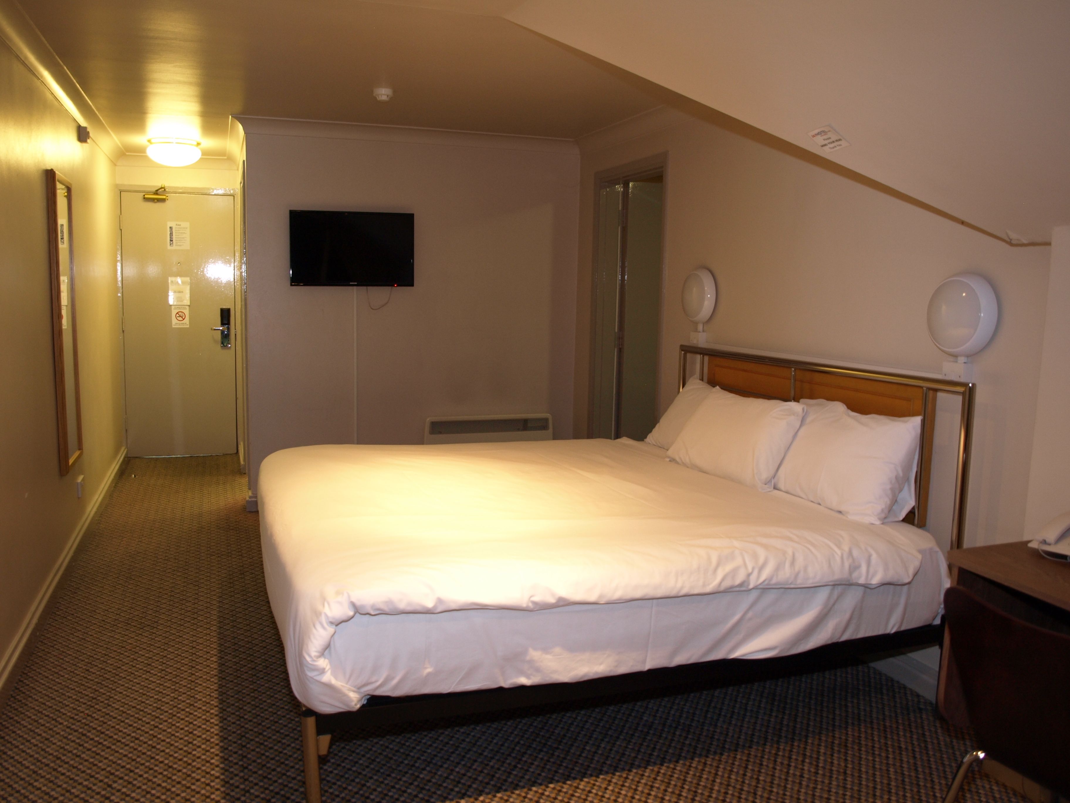 A hotel room with a large bed, a flat-screen TV mounted on the wall, a bedside table, and a mirror.
