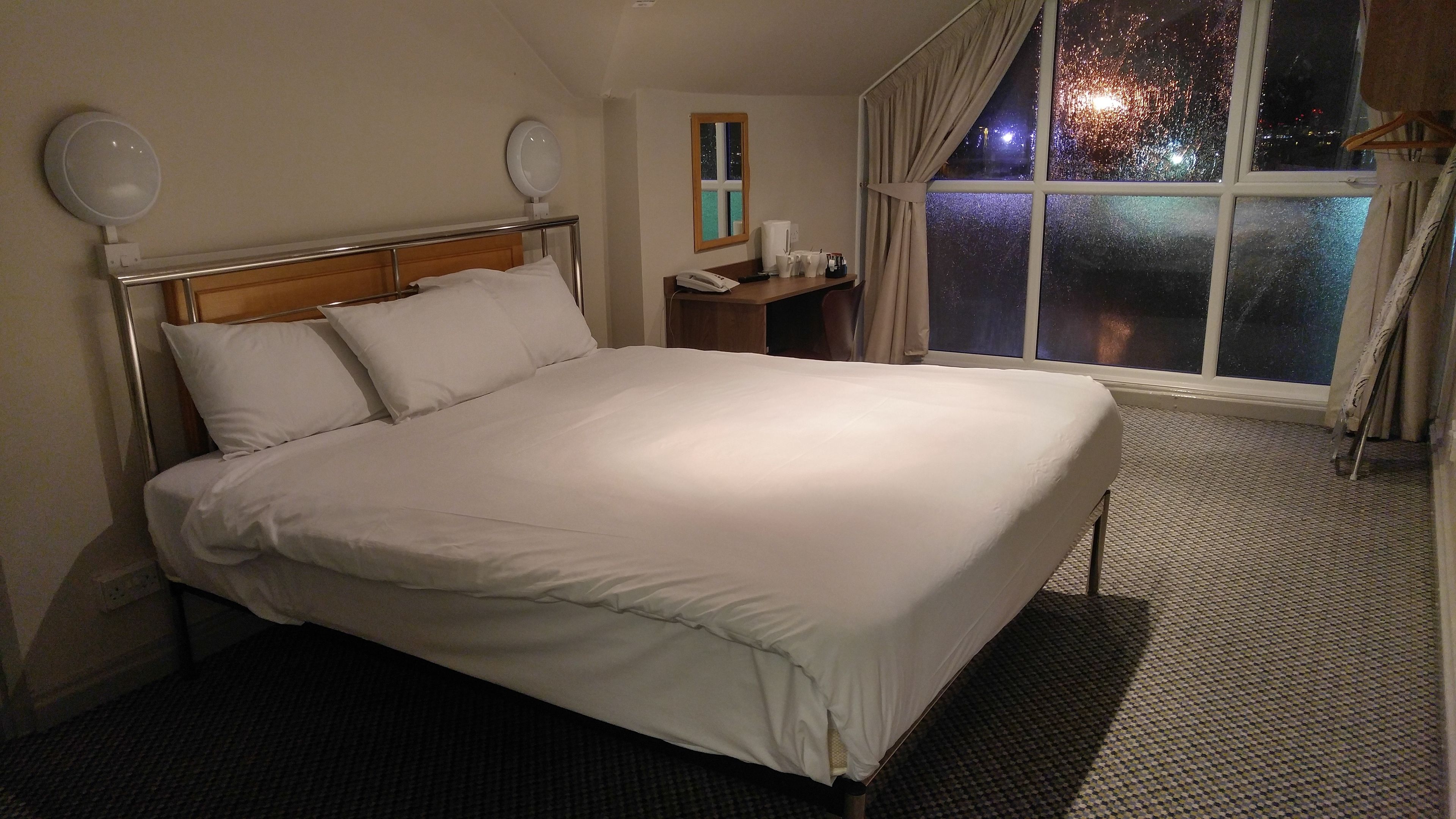 Habitación de hotel con una cama grande, una mesita de noche con un teléfono y una taza, una mesita auxiliar, una ventana con cortinas y un espejo en la pared.