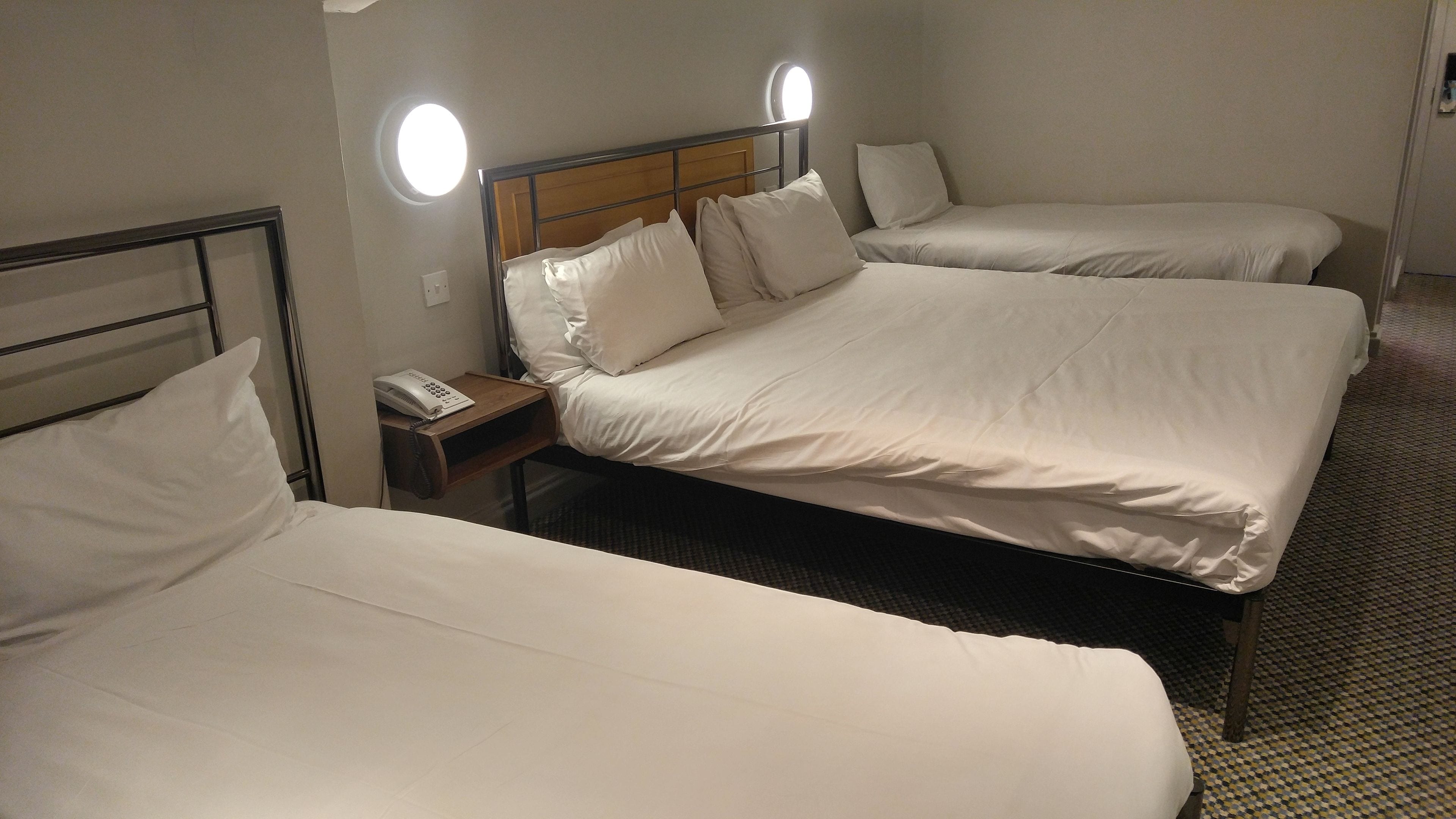 A hotel room with three single beds, each with white bedding and a wooden headboard. There is a bedside table with a phone and a lamp on it.