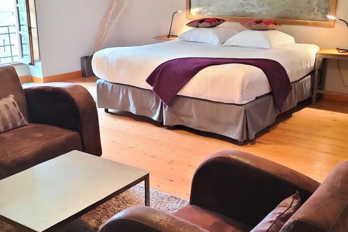 A bedroom with a bed, two armchairs, a coffee table, a painting, and a lamp.