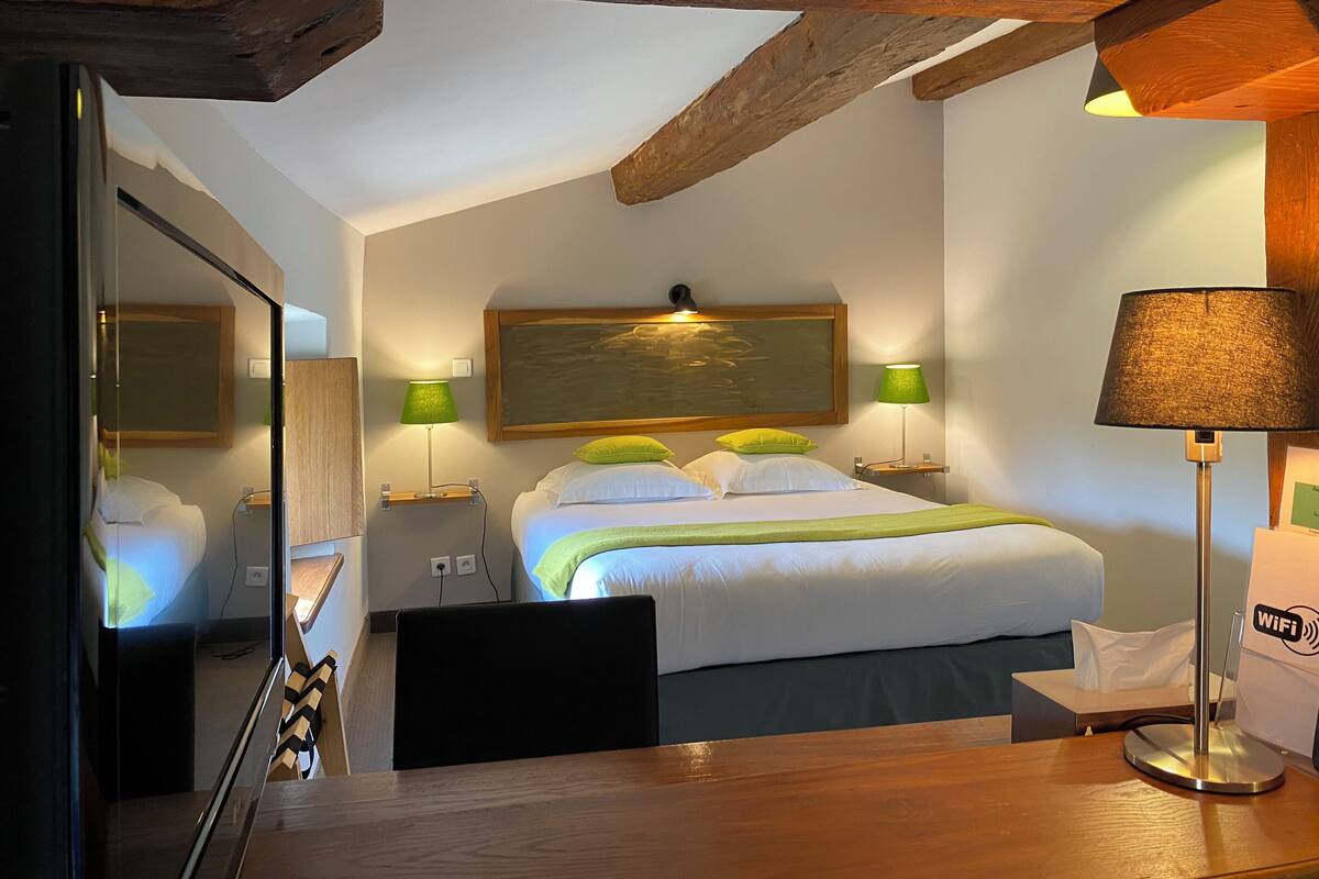 A hotel room with a bed, bedside lamps, a wooden headboard, a mirror, a desk, and a chair.