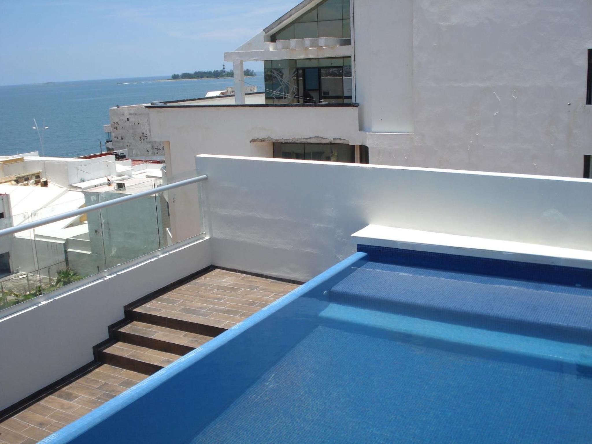 Una piscina en la azotea con barandilla de cristal, vista al mar y un edificio moderno con ventanales amplios.