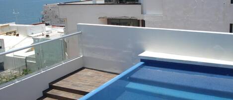 Una piscina en la azotea con barandilla de cristal, vista al mar y un edificio moderno con ventanales amplios.