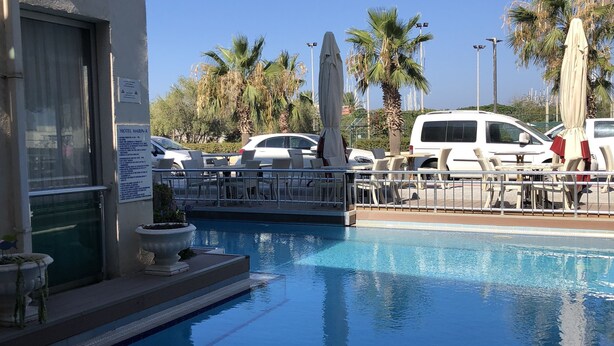 A swimming pool with lounge chairs and umbrellas, surrounded by palm trees and parked cars. - Marin-A Hotel (Bodrum)